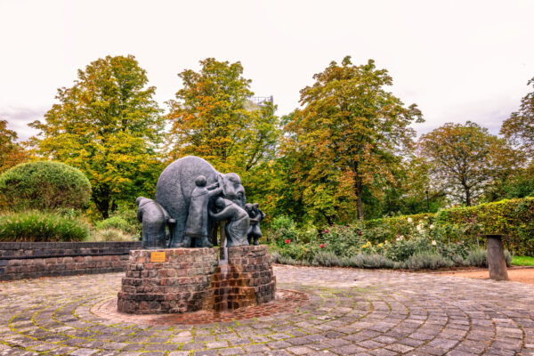 Bronzener Blindenbrunnen in der Bonner Rheinaue. Er zeigt einen Elefanten und mehrere blinde Menschen, die das Tier an verschiedenen K&ouml;rperteilen betasten und so zu unterschiedlichen Schlussfolgerungen gelangen. Eine Informationstafel in Blindenschrift und in normaler Schrift erl&auml;utert den Hintergrund der Darstellung.