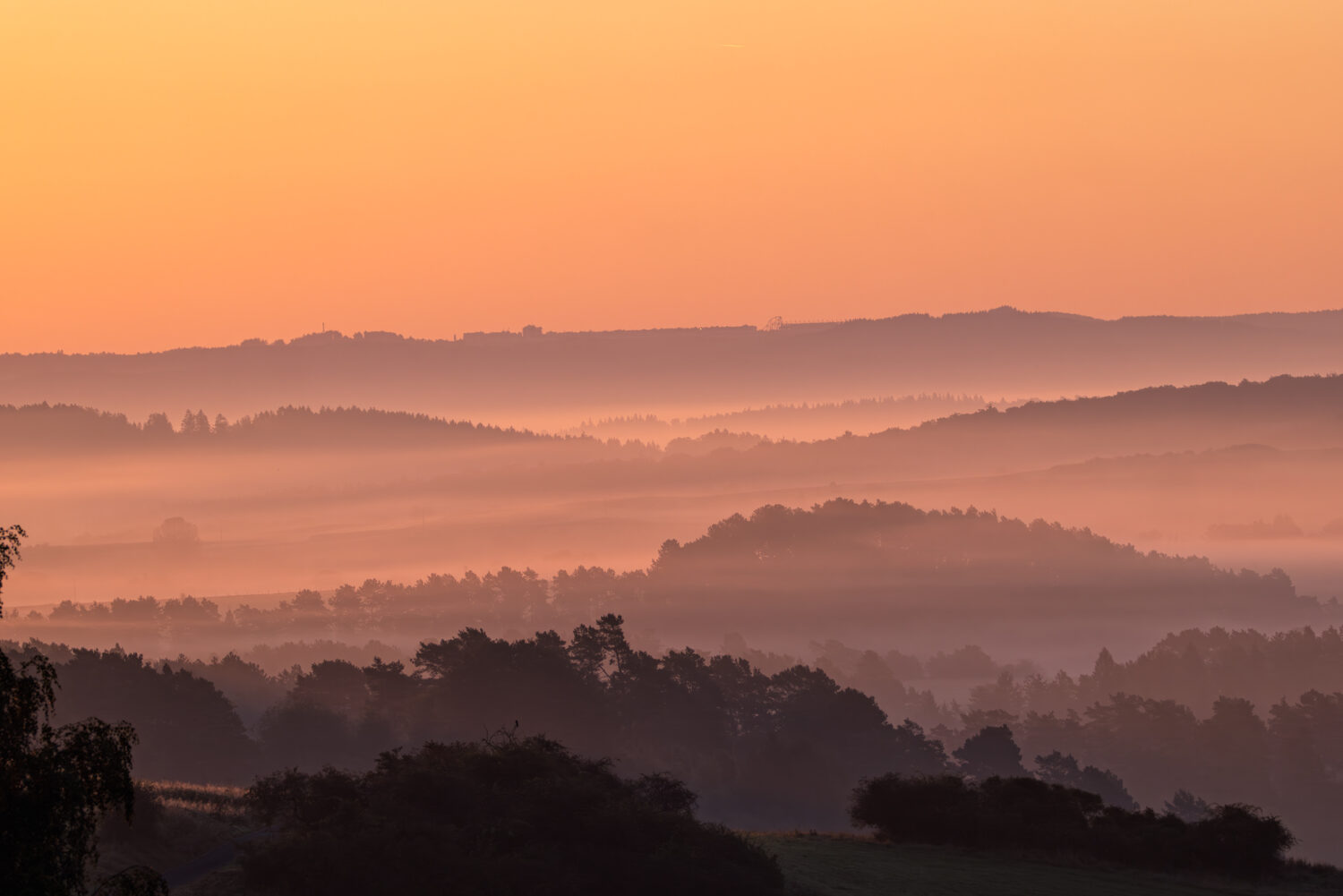 Sonnenaufgang über der Eifel. Im Hintergrund ganz oben erkennt man die Gebäude der Rennstrecke Nürburgring inkl. seiner Achterbahn.