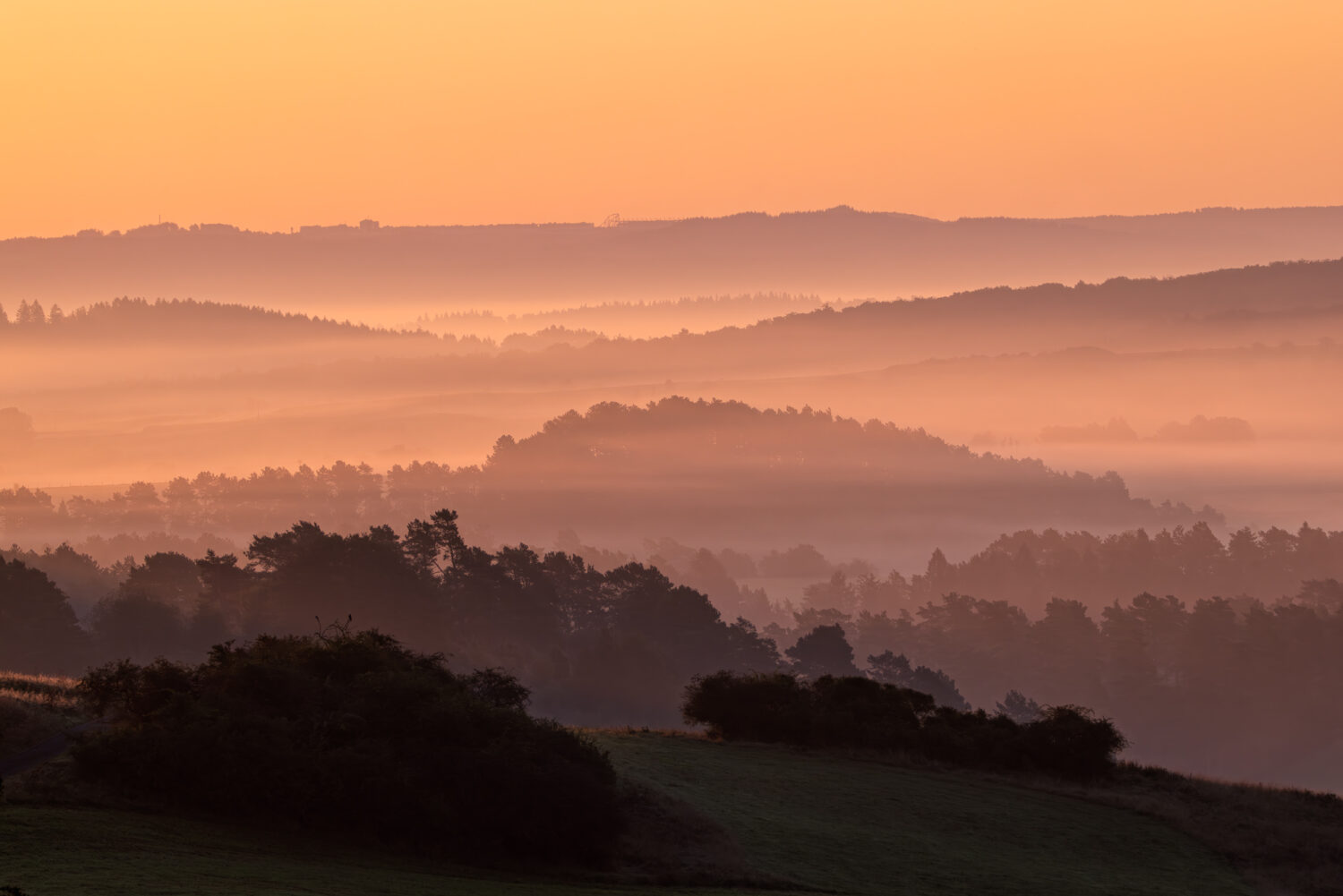 Sonnenaufgang über der Eifel. Im Hintergrund ganz oben erkennt man die Gebäude der Rennstrecke Nürburgring inkl. seiner Achterbahn.